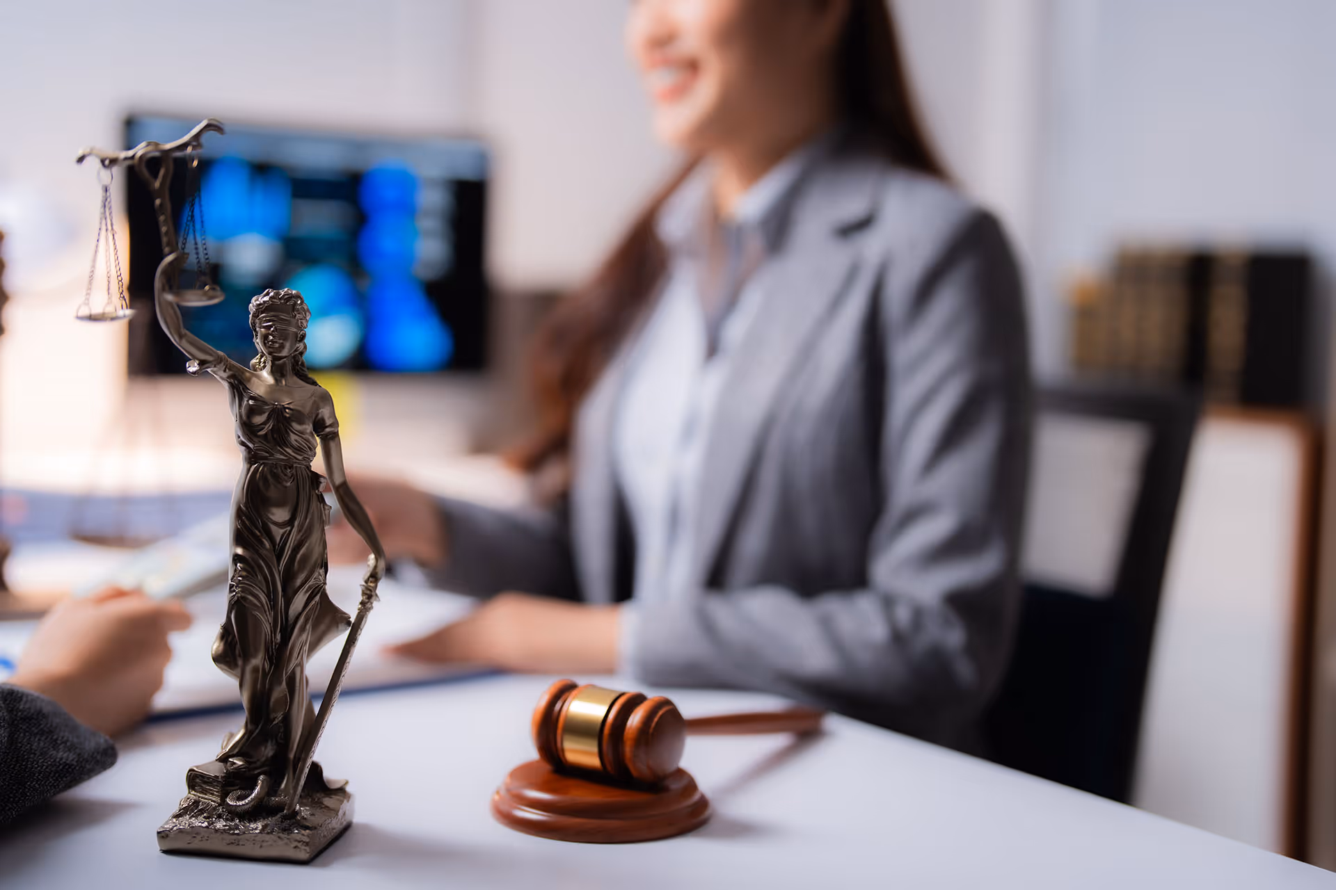 Desk with a Lady Justice statue and a wooden gavel, with a woman in a gray blazer blurred in the background.