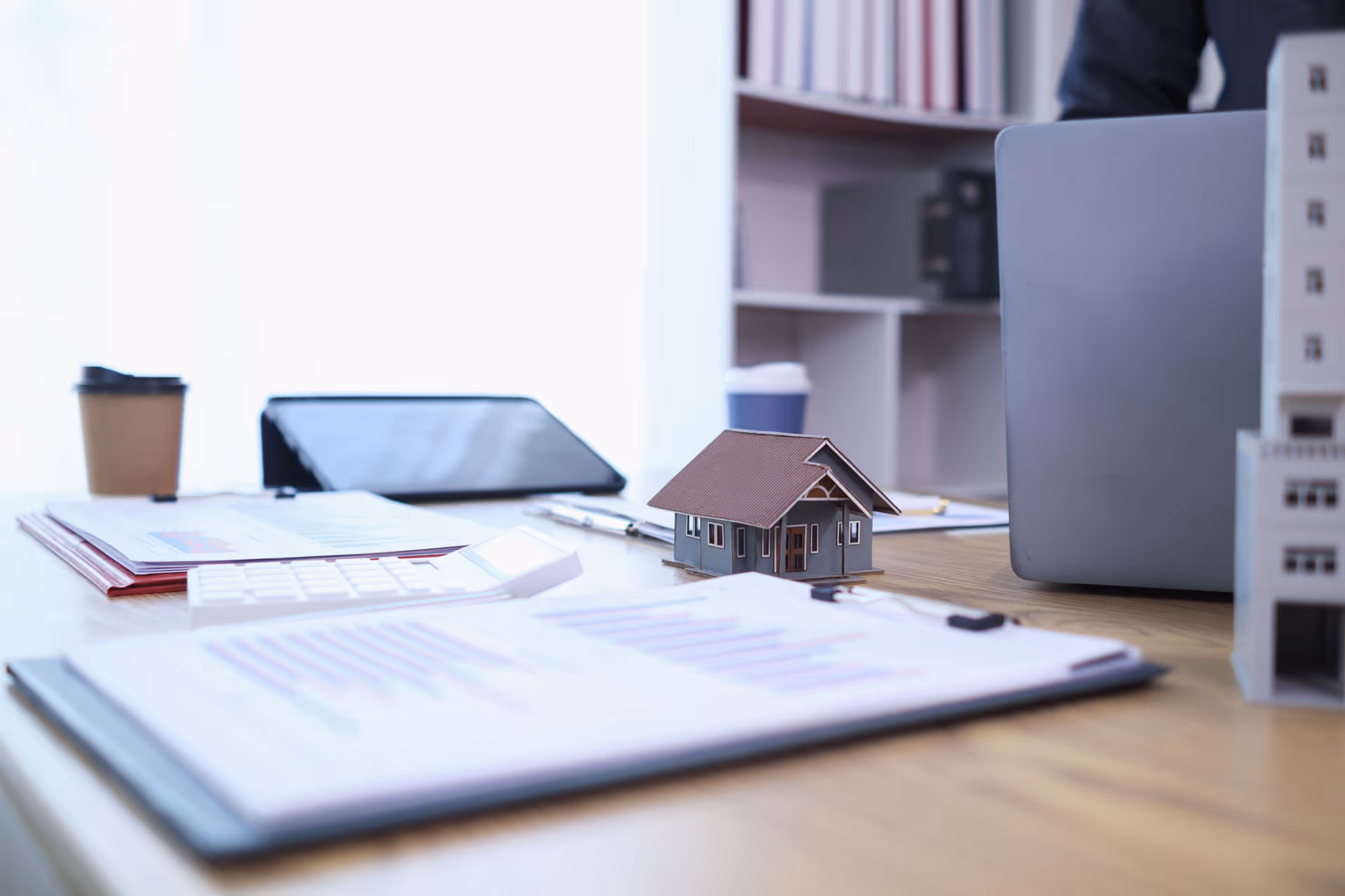 Office desk with a laptop, documents, calculator, coffee cup, and a small house model for real estate planning.