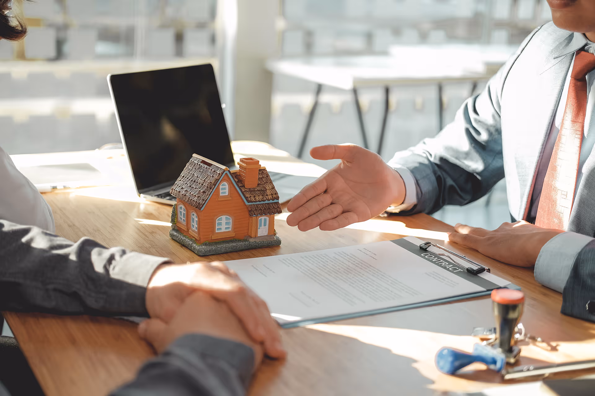 Two people at a desk with a miniature house model and a contract, one person gesturing towards the contract.
