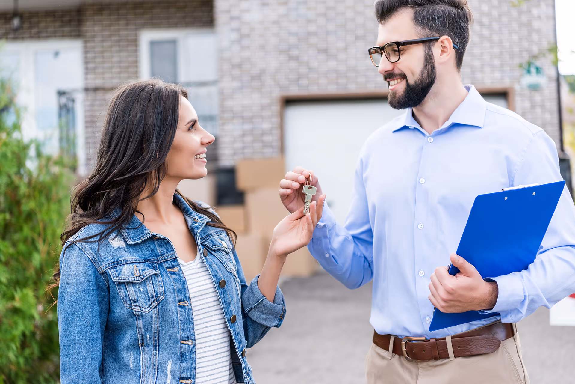 Real estate agent handing keys to a smiling woman standing outside a house with moving boxes in the background.