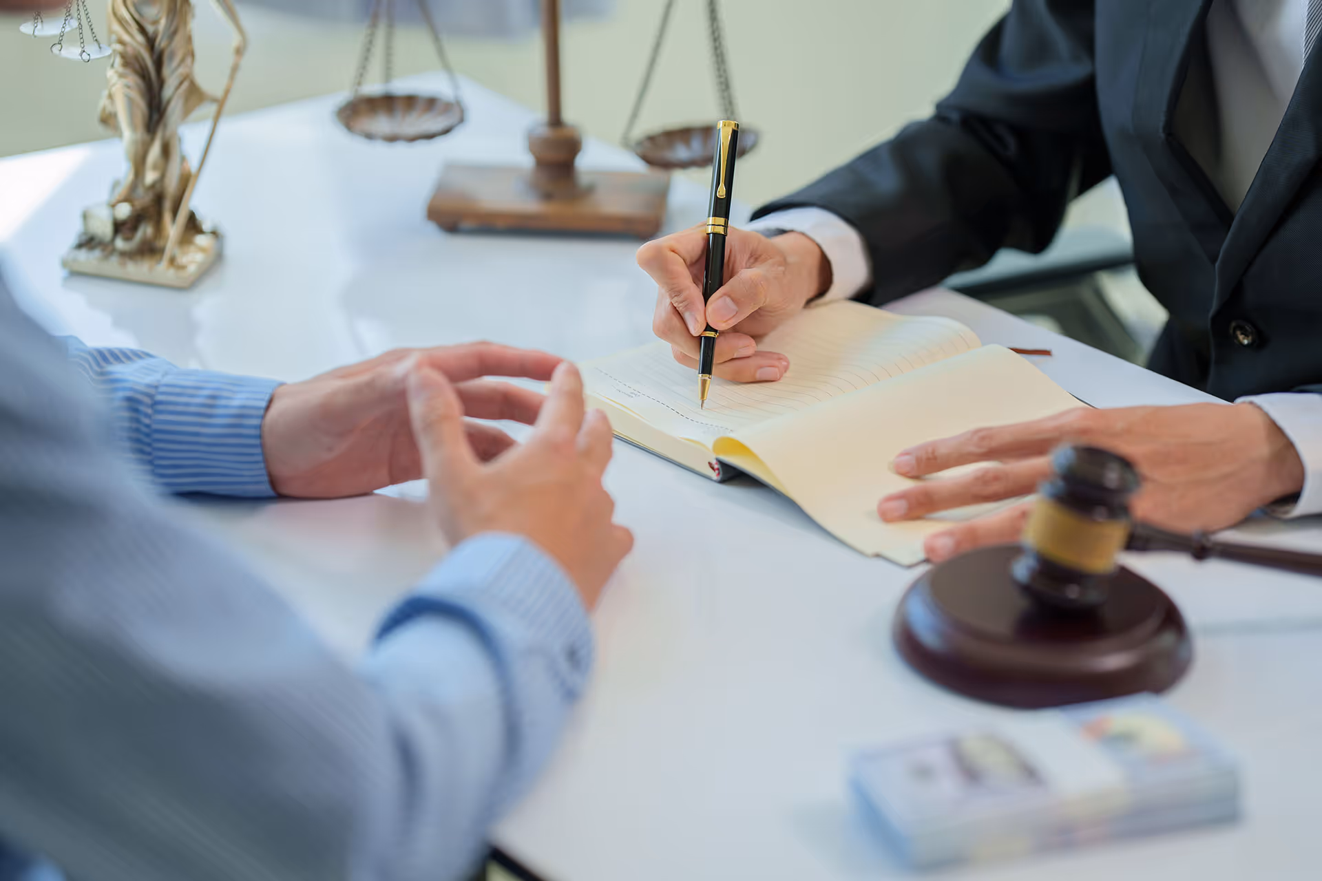 Two people at a desk with legal symbols, one writing in a notebook while the other gestures.