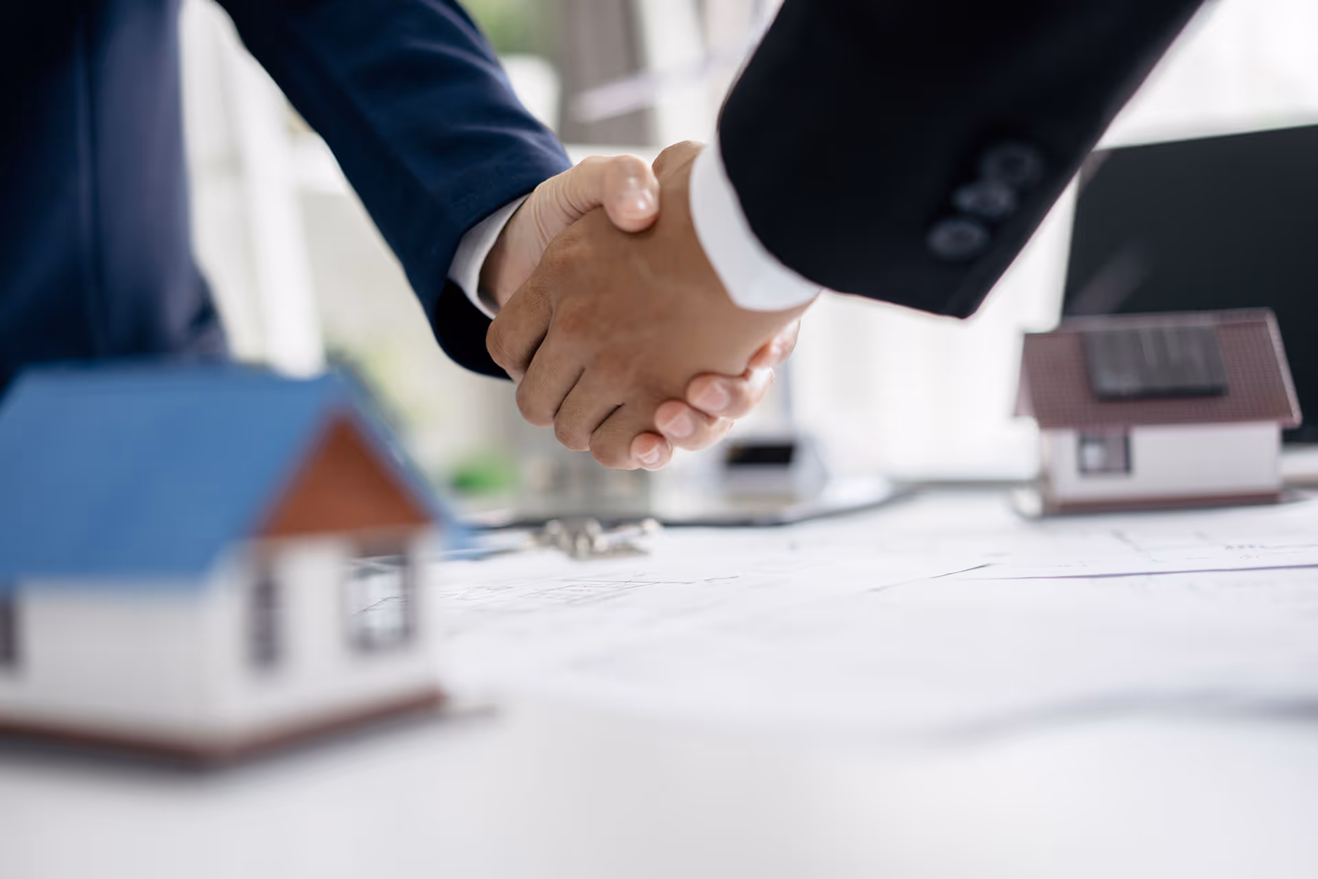 Two businesspeople shaking hands over a table with house models and architectural plans.