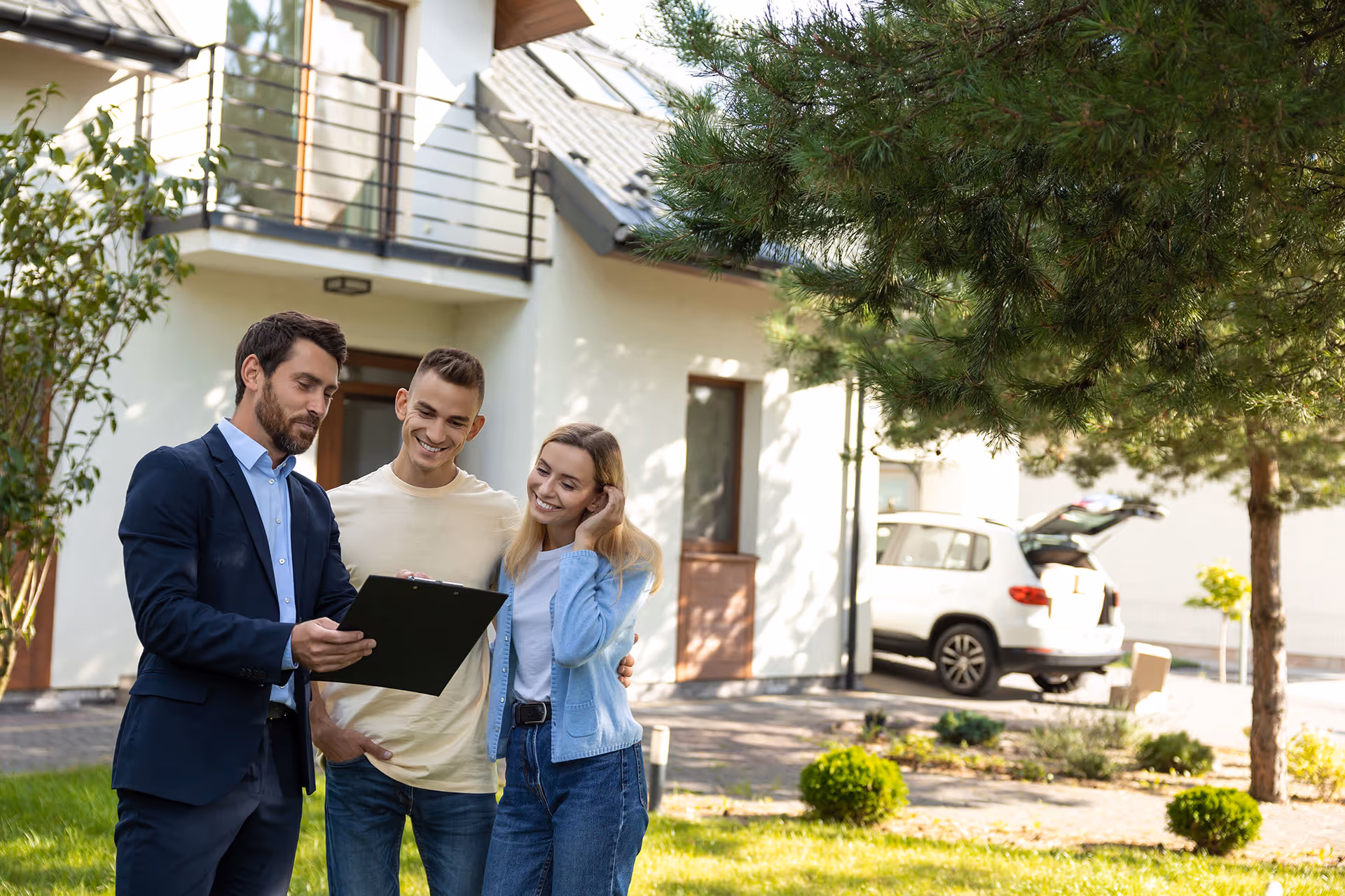 Real estate agent showing a clipboard to a smiling young couple outside a modern house.