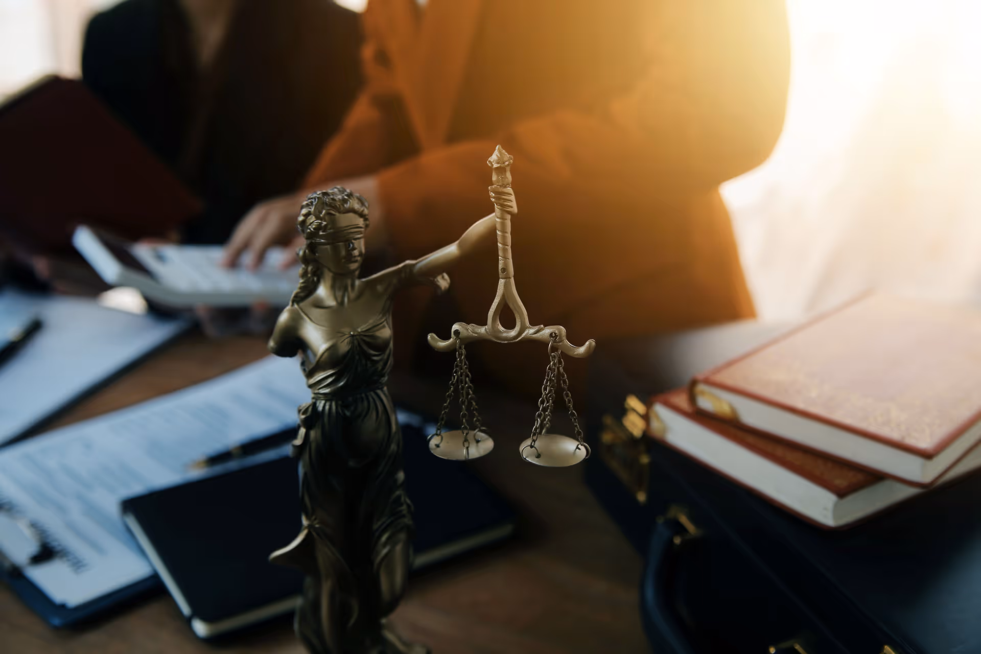 Blindfolded Lady Justice statue holding scales in an office with legal books and documents in the background.
