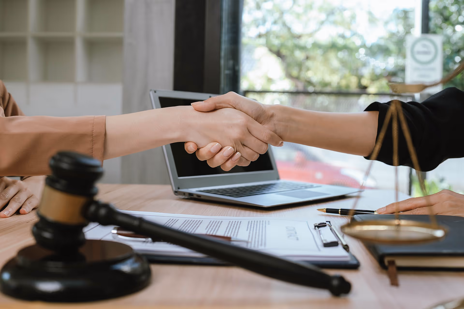 Two people shaking hands over a desk with legal documents, a gavel, and scales of justice nearby.