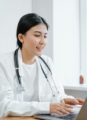 Female doctor wearing a white coat and stethoscope typing on a laptop in a bright room.