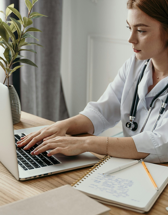 Female doctor in white coat typing on a laptop at a wooden desk with a notebook, pen, and pencil nearby.