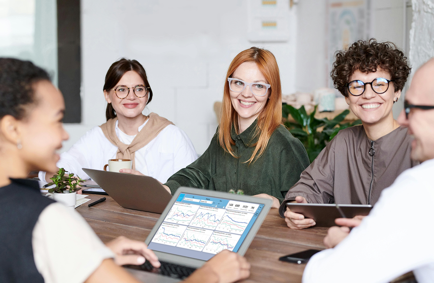 Five diverse people sitting around a table in a meeting, smiling and working on laptops and tablets, displaying data charts on screens.