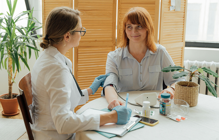 A healthcare professional wearing gloves measures a woman’s blood pressure during a medical checkup in a bright room with plants.