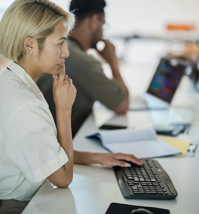 Person with short blonde hair thoughtfully working on a desktop computer keyboard, with an open laptop and documents on the desk.