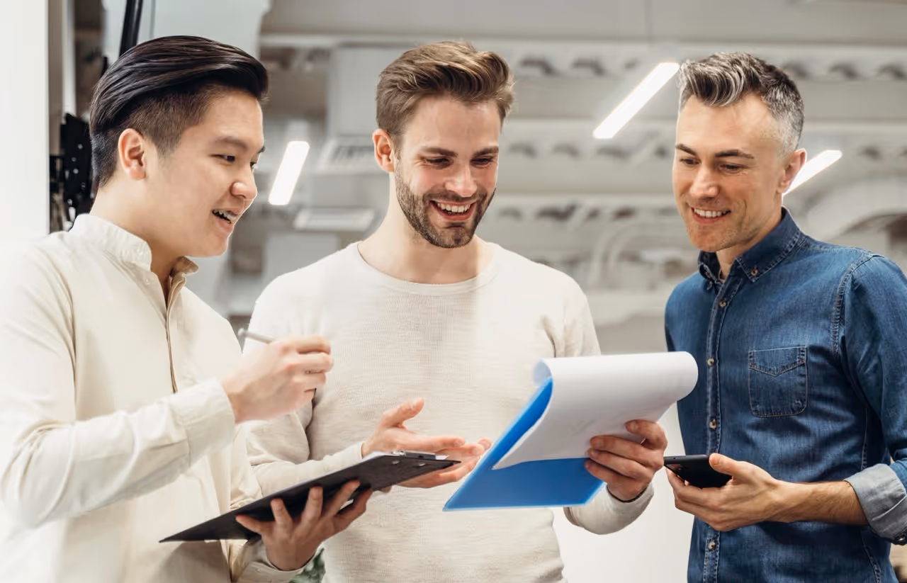 Three men smiling and discussing while reviewing documents and a clipboard in a modern office.