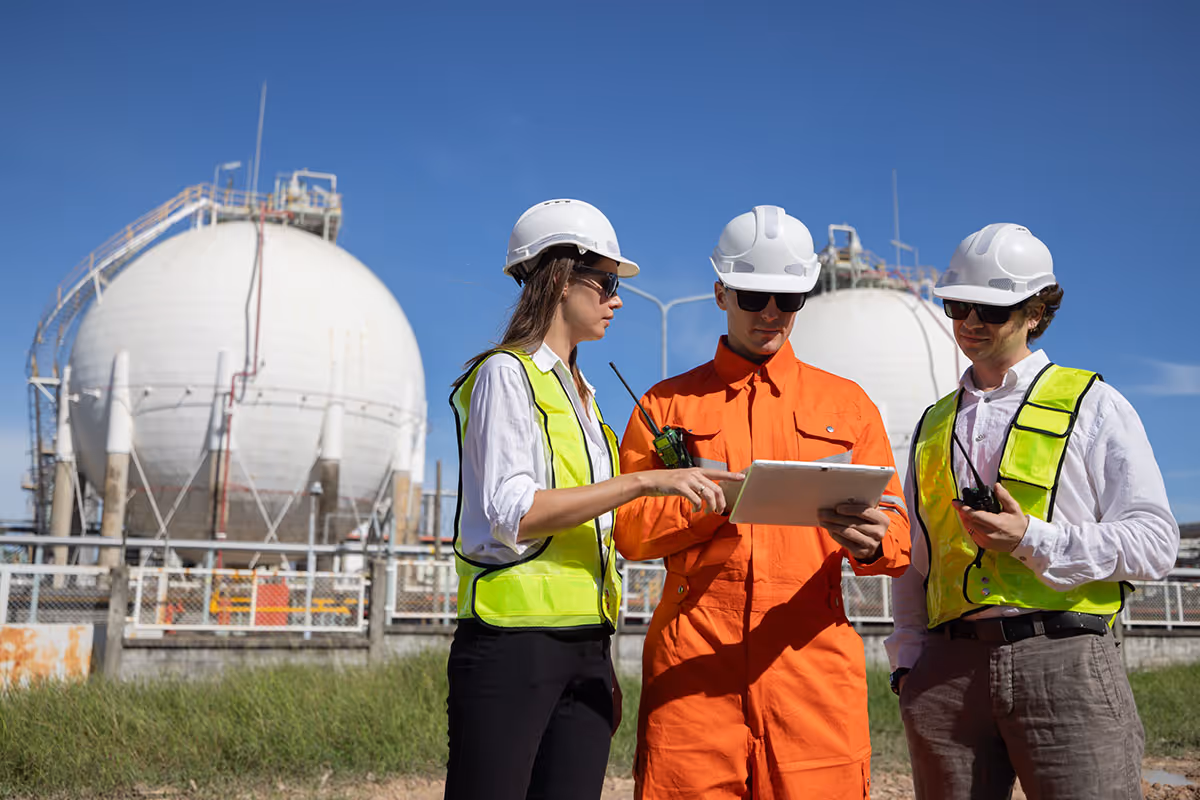Three industrial workers wearing safety helmets and vests reviewing a clipboard in front of large spherical storage tanks at a plant.