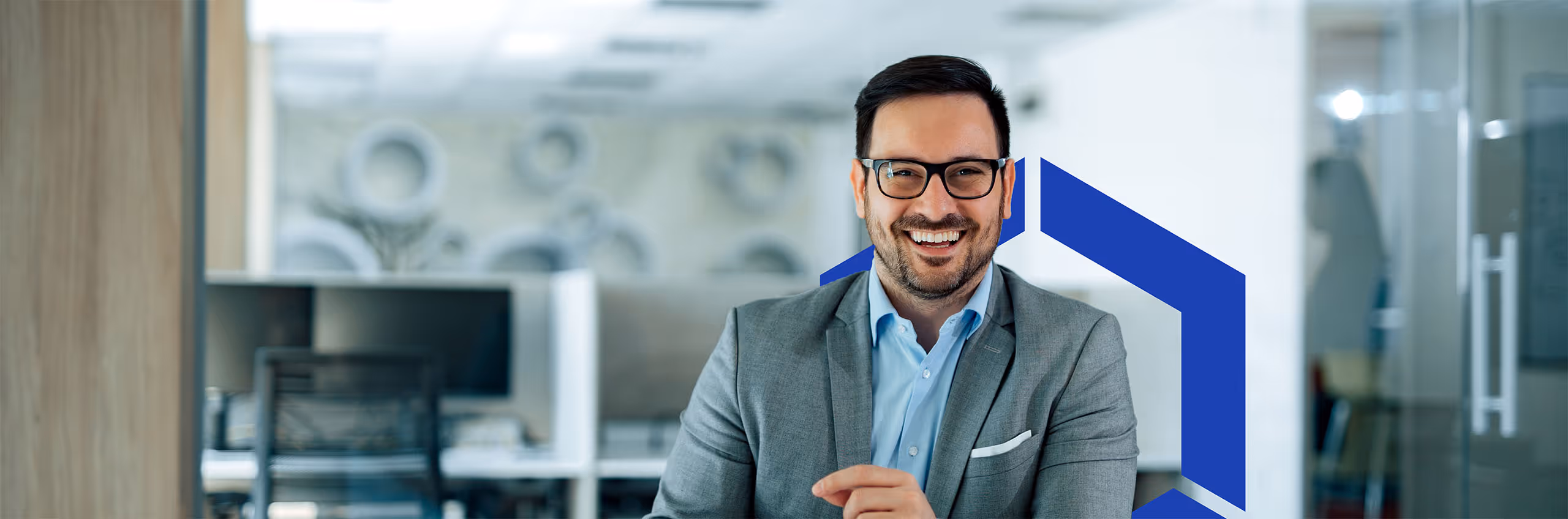 Smiling man in glasses and gray suit sitting in a modern office with a blue geometric design behind him.