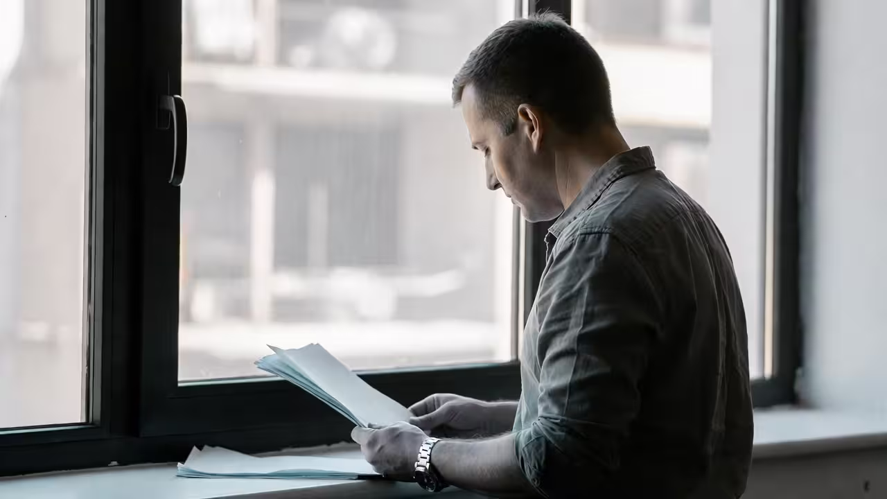 Parent reading a document by a window while managing caregiving responsibilities.