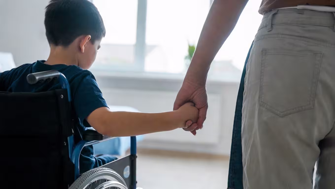 Child in a wheelchair holding a caregiver’s hand for support and guidance.