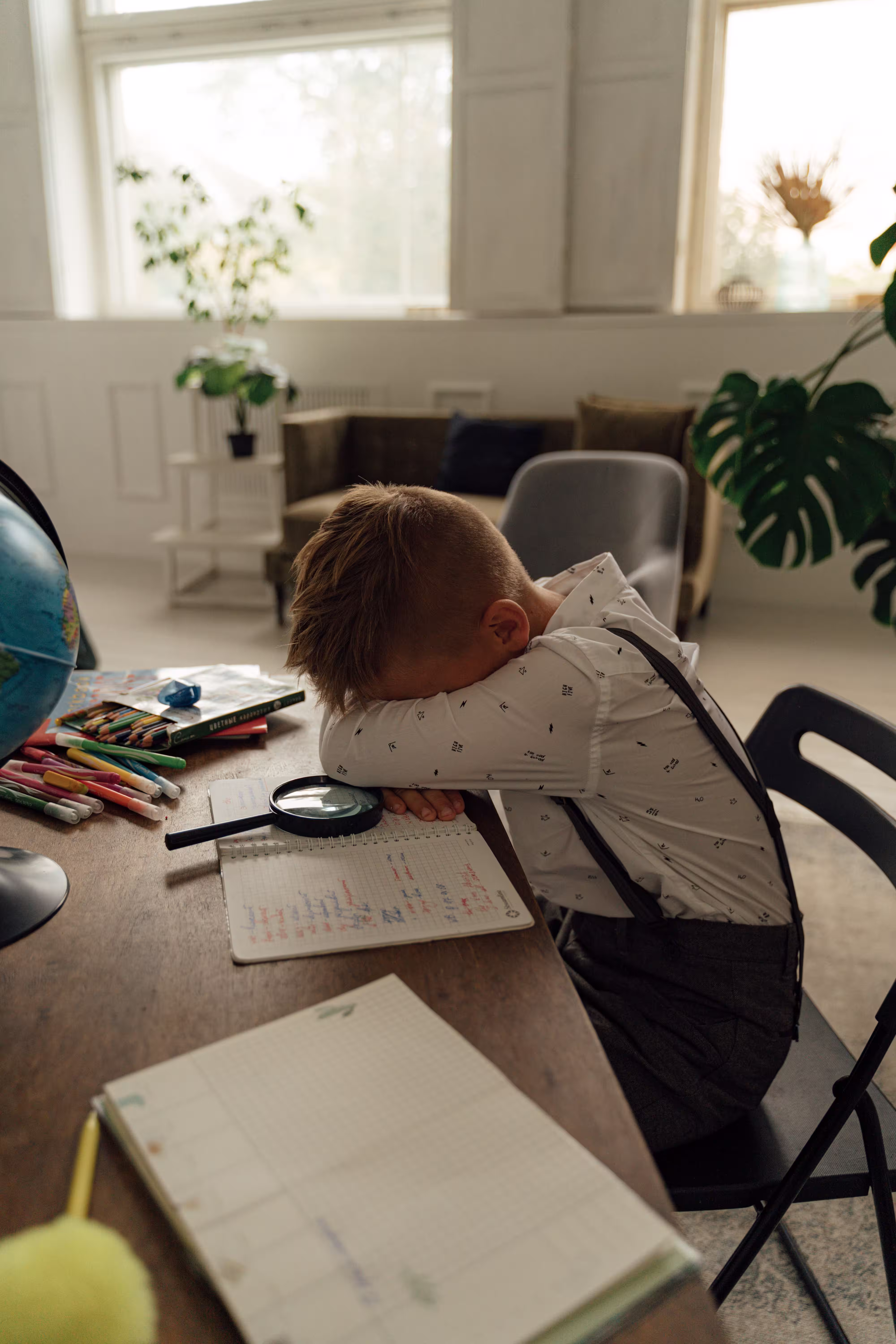 Child resting beside school supplies, colored pencils, books, and a magnifying glass.