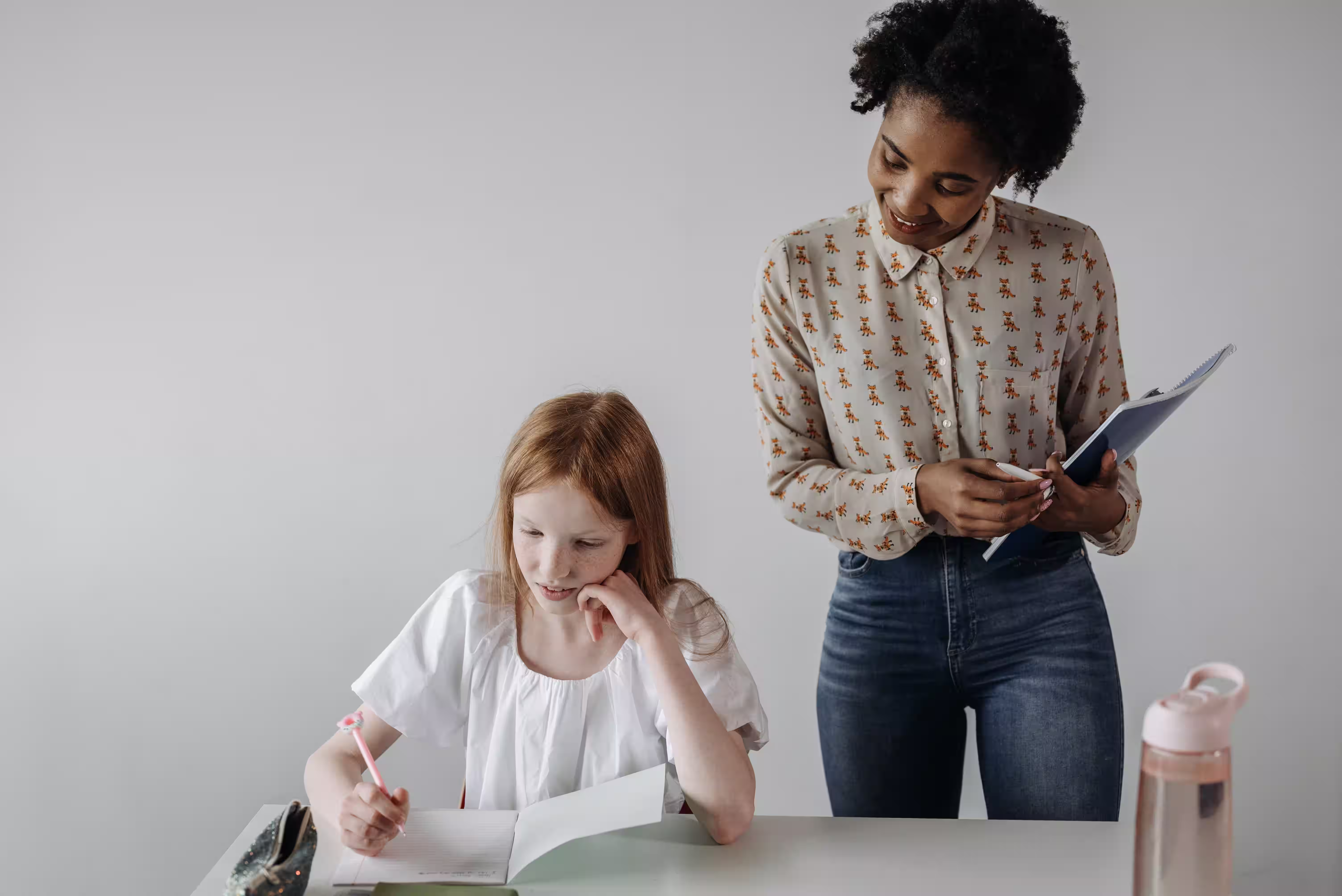 Teacher standing beside a child as they write in a notebook.