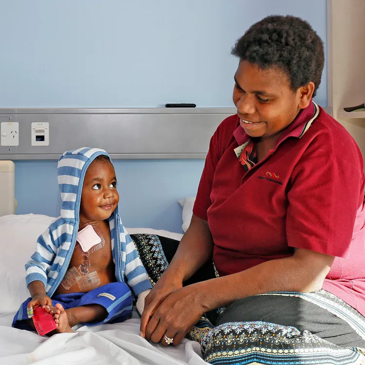 A smiling toddler with medical patches on the chest sits on a hospital bed wearing a blue and white striped hoodie, looking at a woman in a red shirt sitting beside the bed and smiling back.