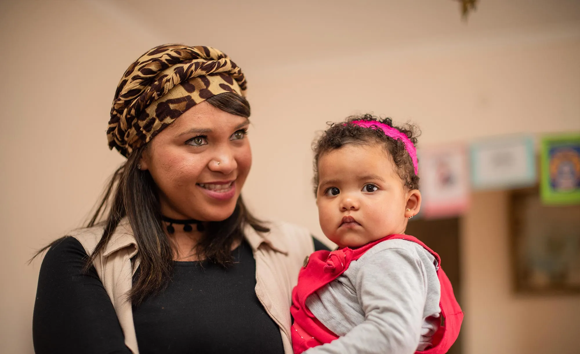 Smiling woman wearing a leopard-print headscarf holding a toddler dressed in a red outfit with a pink headband.
