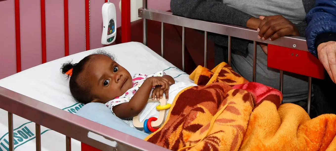 Young child lying in a hospital bed holding a toy, covered with a colorful blanket, with adult hands resting on the bed rail.