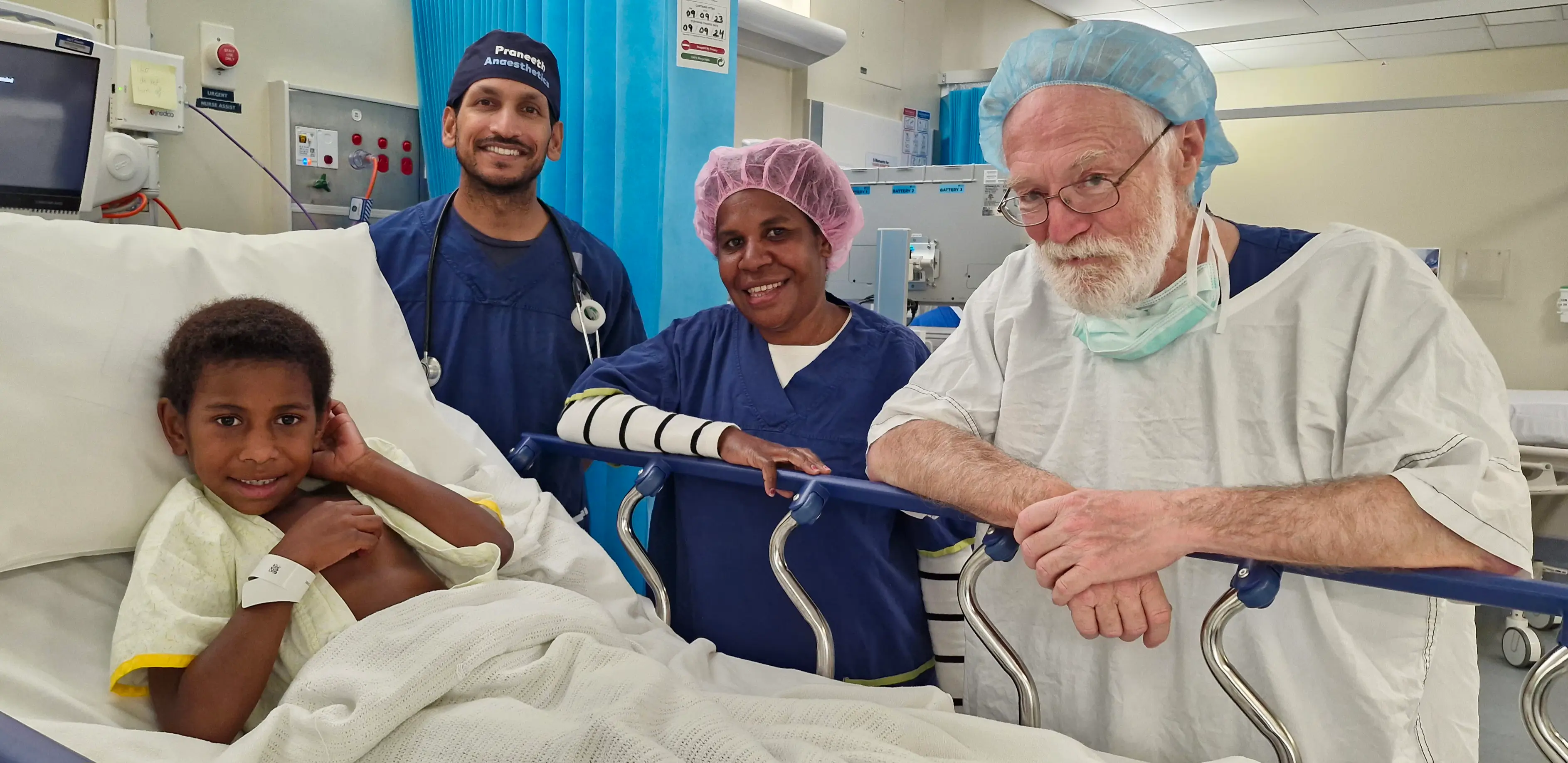 Smiling young patient lying in hospital bed with three healthcare workers in scrubs and surgical caps standing beside him.