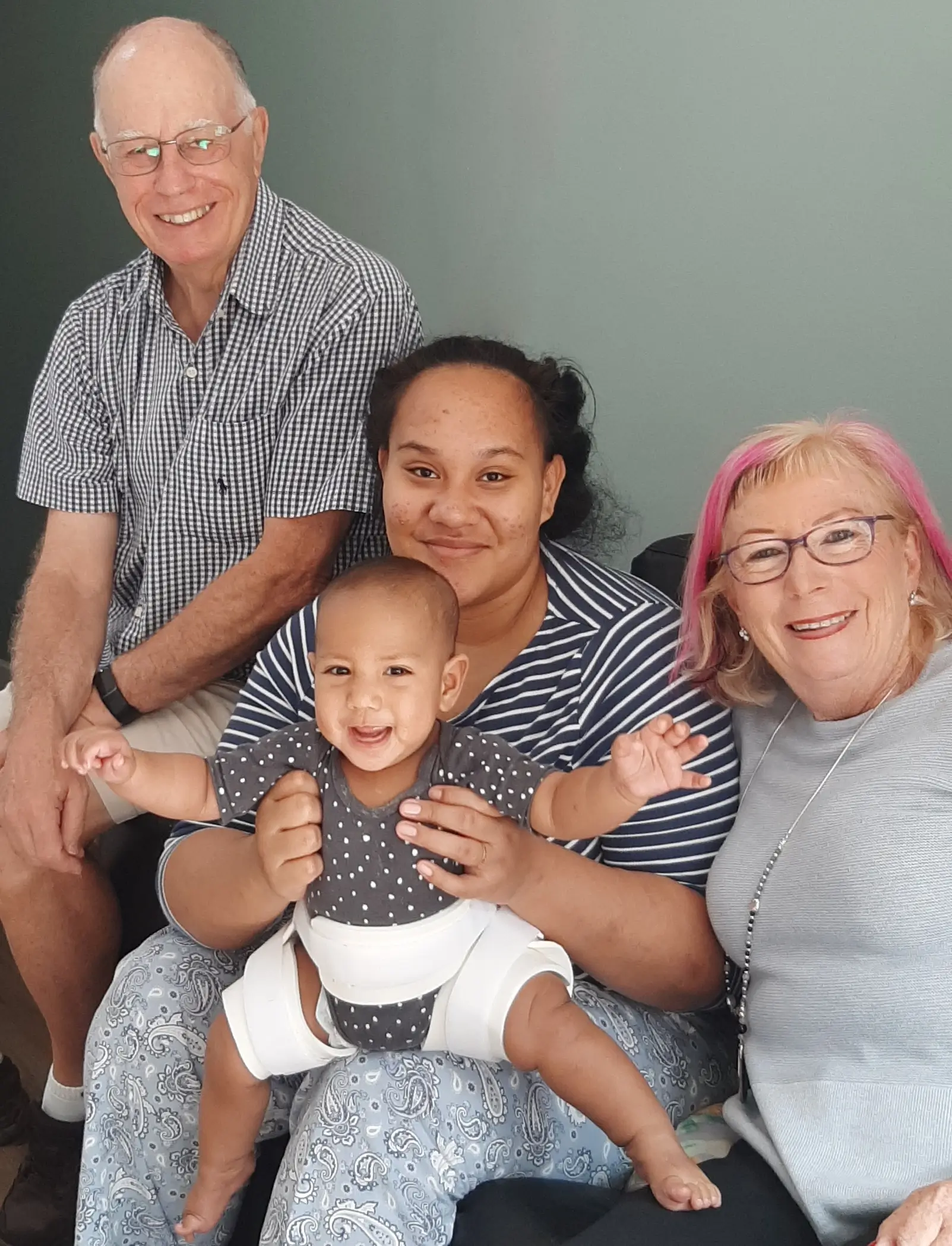 Smiling elderly man, middle-aged woman with baby in leg braces, and older woman with pink highlights sitting together on a couch.