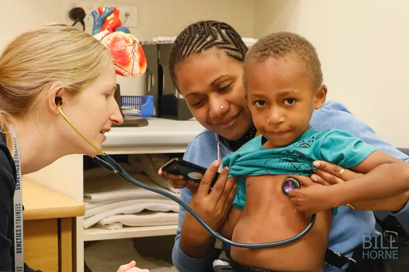 A female doctor uses a stethoscope on a young boy’s chest while his mother supports him and holds a phone.