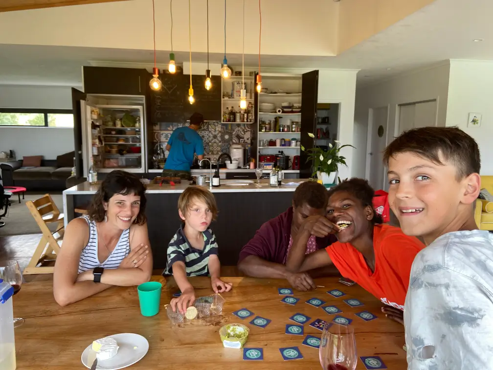 Five people, including children and adults, sitting around a wooden table playing a card game and eating snacks in a modern kitchen.