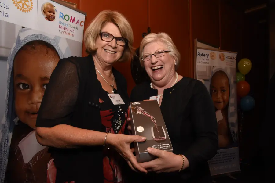 Two women smiling and holding a boxed aerating pourer gift at a Rotary Oceania Medical Aid for Children event.