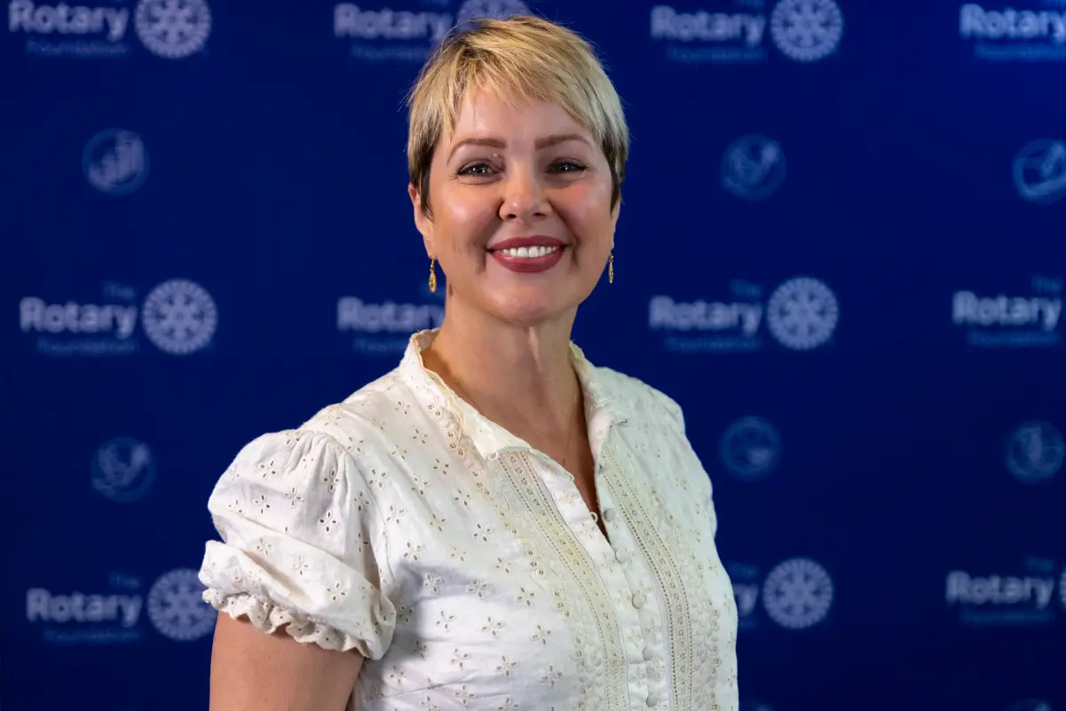Smiling woman with short blonde hair wearing a white blouse stands in front of a blue backdrop with Rotary Foundation logos.