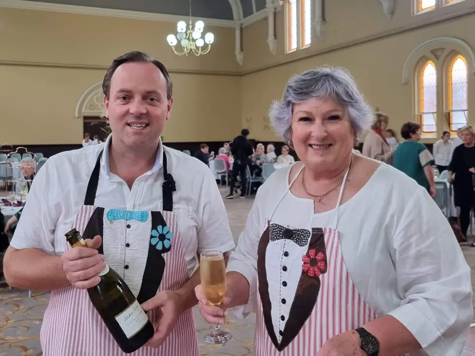 Smiling man and woman wearing aprons designed like tuxedos, holding a bottle of champagne and a glass in a banquet hall.