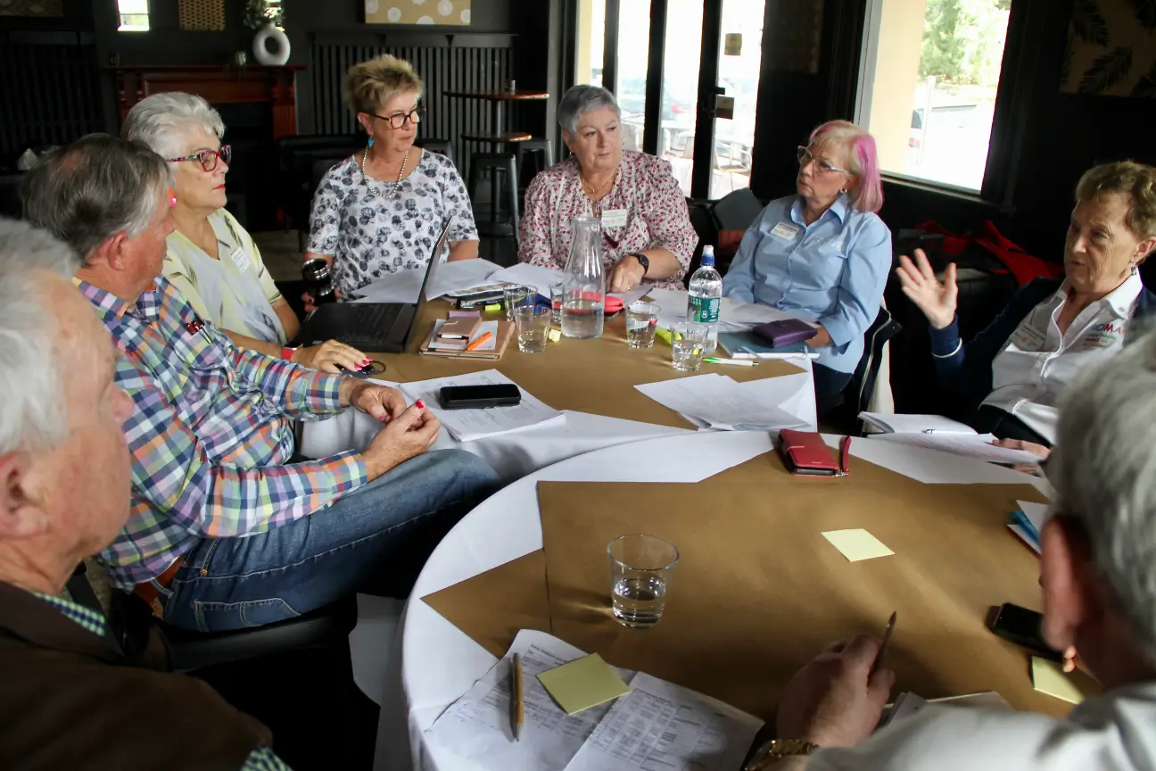 Group of older adults engaged in a discussion around a table with papers, notebooks, and glasses of water.