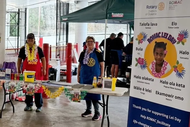 Two women wearing leis and aprons standing behind a decorated fundraising table at an outdoor event with a ROMAC Lei Day banner featuring a girl and greetings in Pacific Island languages.