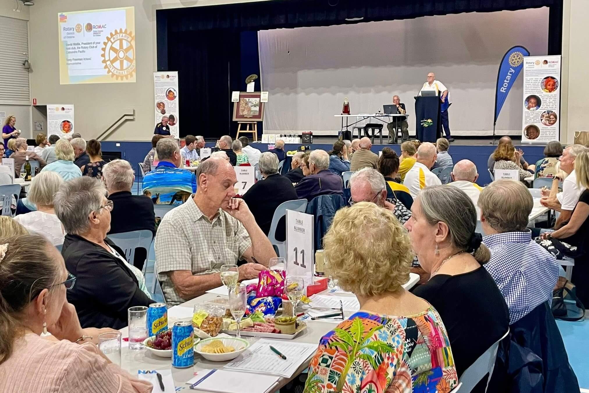Adults seated at tables with snacks and drinks attending a Rotary Club trivia night event in a hall with a speaker at a podium on stage.