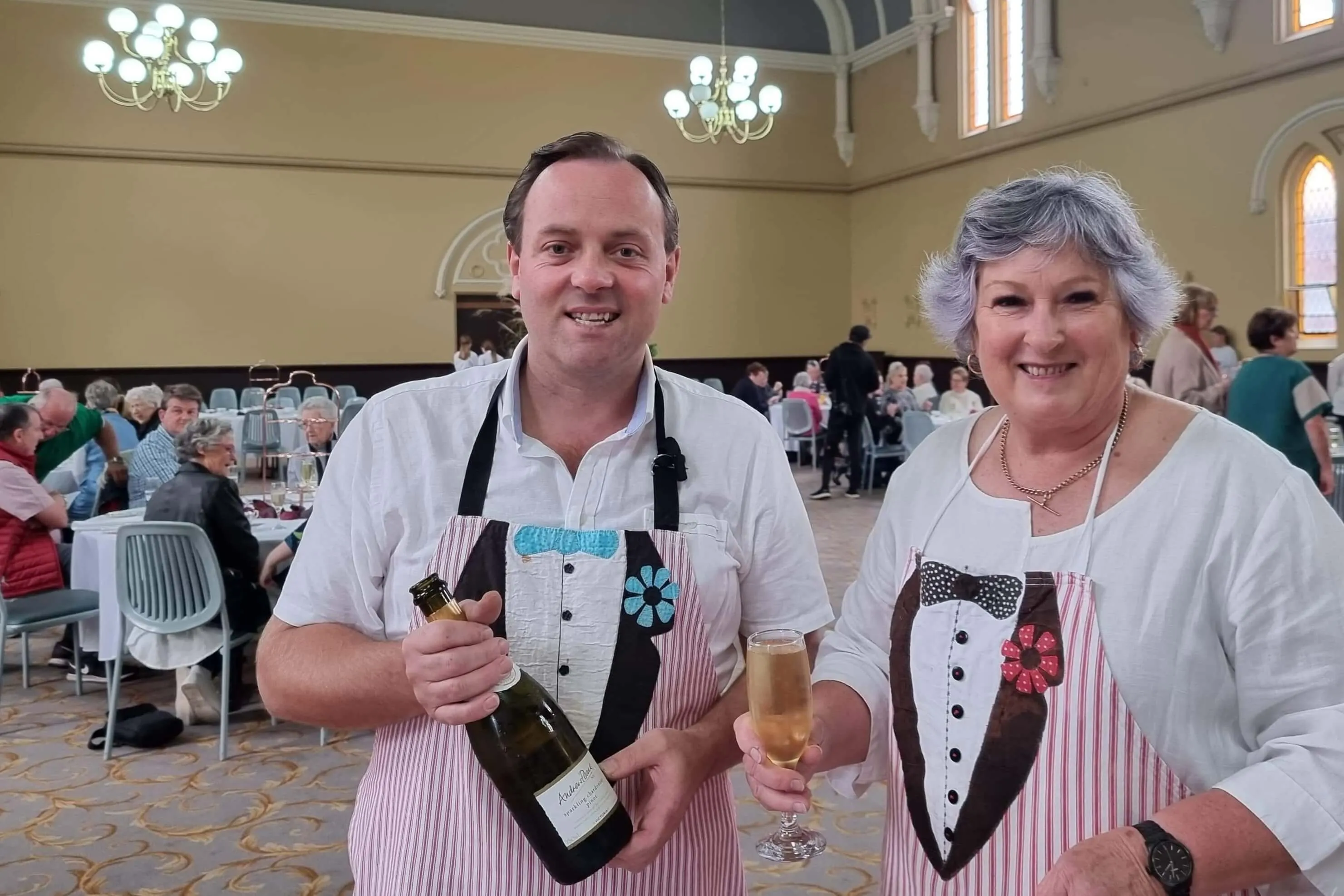 Man holding a bottle of sparkling wine and woman holding a glass of sparkling wine, both wearing aprons with tuxedo designs, smiling indoors at a social gathering.