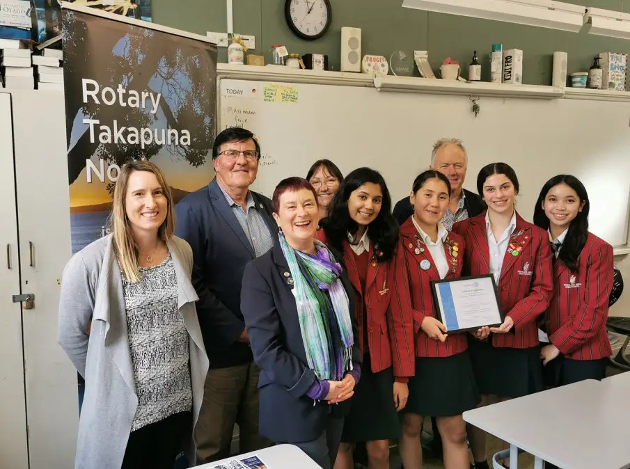 Group of seven people, including four girls in red striped school blazers holding a certificate, standing in a classroom with a Rotary Takapuna banner in the background.