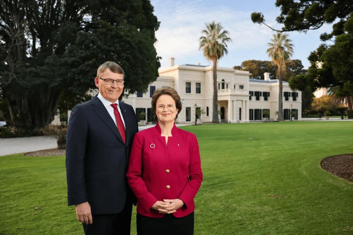 A man in a dark suit and red tie stands next to a woman wearing glasses and a red jacket, both smiling in front of a large white building with palm trees and green lawn.