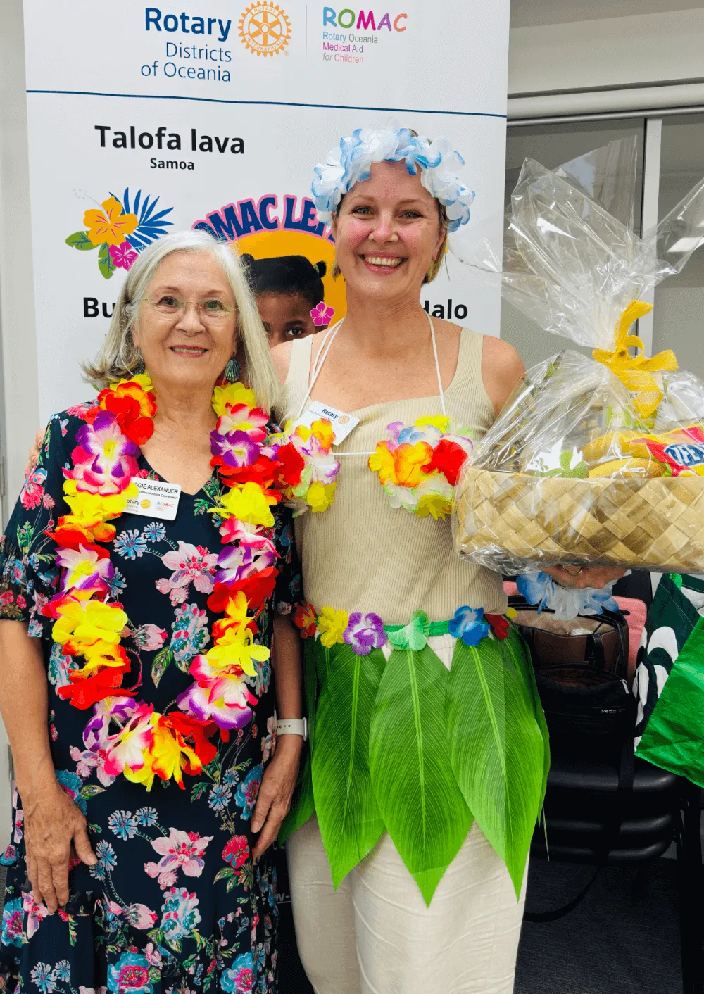 Two smiling women wearing colorful flower leis and tropical attire, with one holding a gift basket in front of a Rotary Oceania banner.