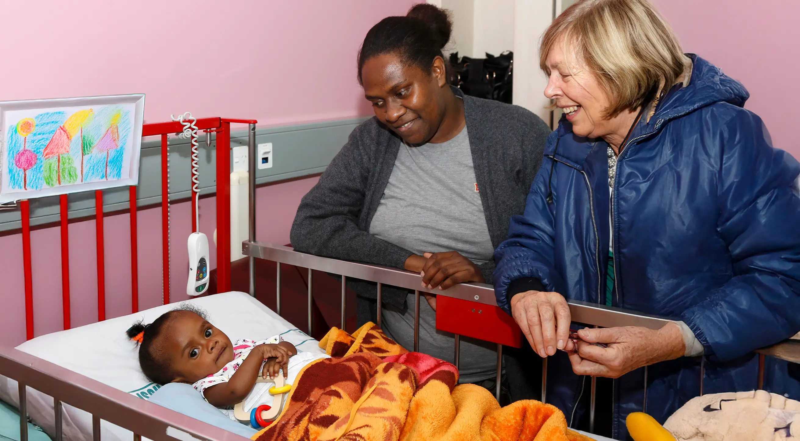 A baby lying in a hospital crib with a colorful blanket, watched by a woman in a gray sweater and a woman in a blue jacket smiling.