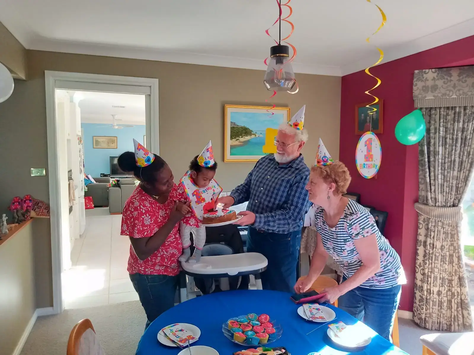 Family celebrating a baby's first birthday with party hats and a birthday cake with a candle in a decorated room.