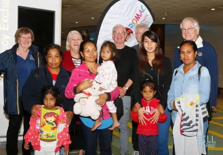 Group photo of diverse adults and children standing indoors, some holding younger children, with a ROMAC banner in the background.