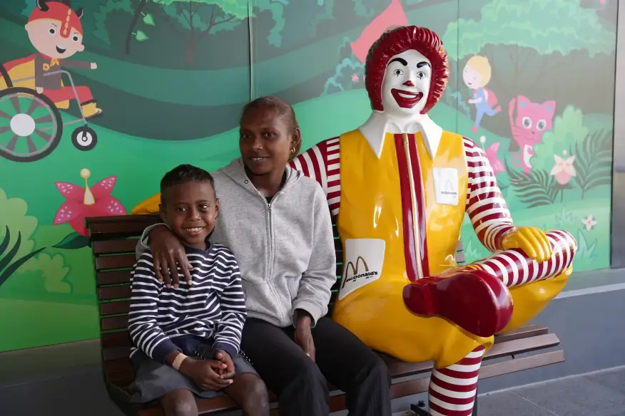 A woman and a child sitting on a bench next to a Ronald McDonald statue, with a colorful mural in the background.