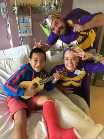 Smiling boy with a red leg cast playing a small guitar in a hospital bed, accompanied by two adults in purple and yellow superhero costumes making playful gestures.