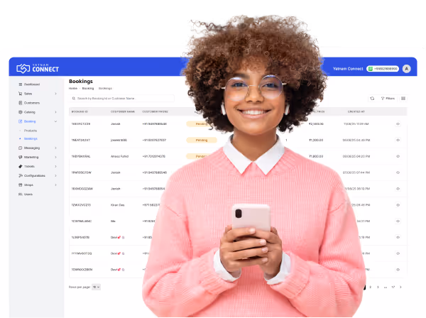 Smiling woman with curly hair and glasses wearing a pink sweater, holding a smartphone in front of a Yatnan Connect bookings dashboard screen.