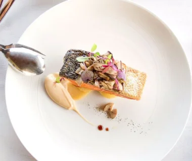 Finely prepared dish: a rectangular, fried fish fillet with crispy skin on a slice of toasted bread, garnished with sautéed mushrooms and microherbs; next to it a creamy sauce, a few small dark sauce dots and a spoon on the edge of the plate on a large white porcelain plate, bright, elegant fine dining atmosphere.
