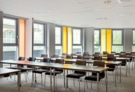 Empty modern seminar or conference room with several rows of simple wooden tables and black chairs, large windows with yellow window panels, bright daylight flows in and offers views of surrounding buildings and trees; bright acoustic ceiling, grey carpet and quiet, tidy atmosphere.