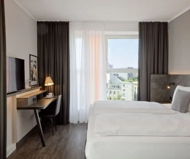 Bright, modern hotel room with two single beds standing next to each other with white sheets and gray pillows; large window with white curtains and dark curtains offers natural light and views of neighboring buildings. On the right, an upholstered headboard with a wall-mounted reading lamp, on the left, a narrow wooden desk with chair, table lamp, teapot and wall-mounted flat screen; dark wooden floor, quiet and minimalistic atmosphere.