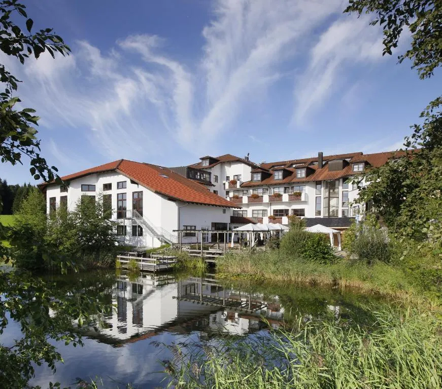 Large white hotel with red tiled roofs and several floors on the banks of a quiet pond; facades with balconies and flower boxes, terrace with parasols and a wooden walkway, everything is reflected in the water. Surrounded by reeds, banks and trees, with meadows and a blue sky with feathery clouds in the background. Mood: peaceful and rural.