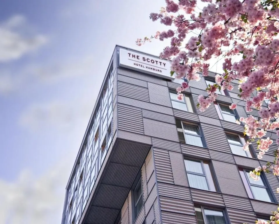 Lower perspective of a modern six-story hotel with grey metal facades and regularly arranged windows; at the top a sign with the inscription “THE SCOTTY HOTEL HAMBURG”. In the foreground on the right, a branch with pink cherry blossoms, blue sky with soft clouds in the background. Mood: spring-like, urban and inviting.