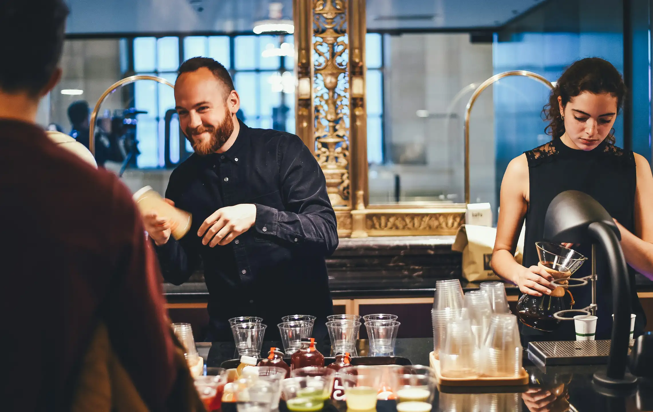Two baristas behind a café counter, in the foreground a customer only visible with their backs. On the left, a smiling, bearded man in a dark shirt handing a mug to a guest; on the right, a concentrated young woman pouring hot filter coffee into a glass carafe with a wooden neck. On the counter, several plastic cups, syrup bottles and small sample bowls; in the background, an ornate golden mirror frame and warm, inviting light. Mood: friendly and lively.